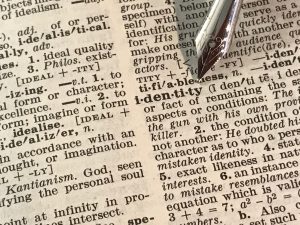 a quill tip points to the word "identity" on the page of an open dictionary