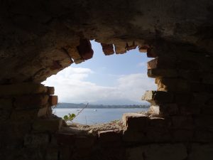 a large hole in a brick wall foreground reveals a large lake with sailboats in the background