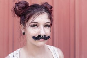 a freckled young woman smiles wearing a fake mustache and nose piercing