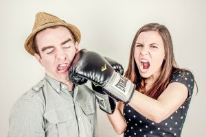 an angry woman wearing boxing gloves punches a hat-wearing man in the jaw