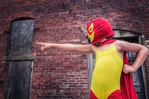 A child wearing a leotard, cape, and a mask has one fist on their hip and the other arm extended with a pointing index finger