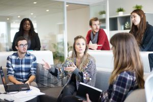 a group of people are gathered in a casual office setting. a white woman in the center explains something to the mostly white group, while a Black woman in the background looks on