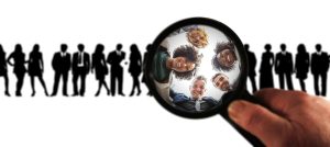 a hand holding a magnifying glass over a lineup of business professionals' silhouettes. inside the magnifying glass, we see a circle of five smiling faces