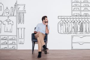 Dreaming about new wardrobe. Young man keeping hand on chin and looking away while sitting in the chair against illustration of closet in the background