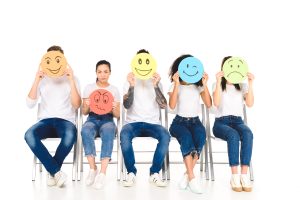 five people representing a variety of genders and ethnicities are seated in a row of chairs. each holds a different cardboard emoji over their face, except for one woman who holds a frustrated emoji in front of her torso