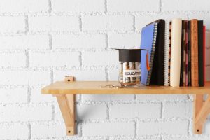 Glass jar with money for education, books and notepads on wooden shelf