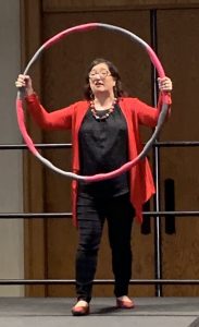 Amy Waninger stands on a stage, holding up a hula hoop with both hands